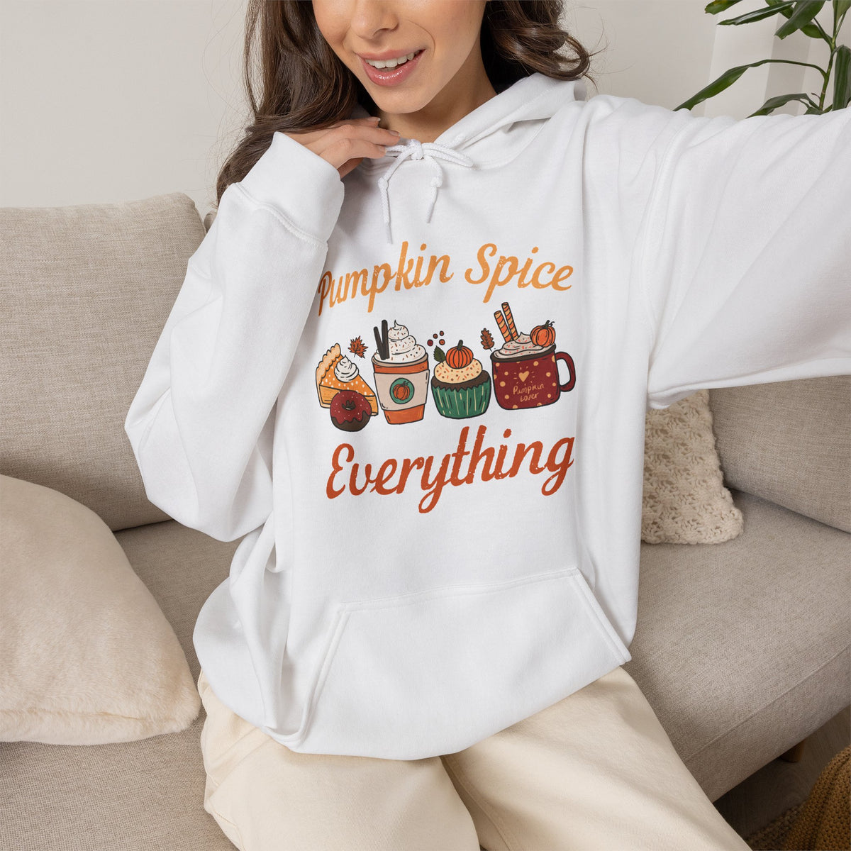A woman wearing a white pullover hoodie with a colourful autumn-themed graphic that reads 'Pumpkin Spice Everything' and shows coffee treats, sitting on a beige couch.