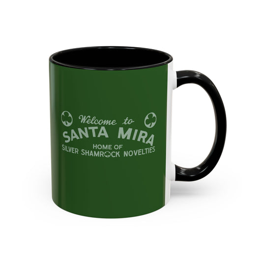 A ceramic coffee mug with a dark green exterior, black interior and handle, and white printed text reading 'Welcome to SANTA MIRA HOME OF SILVER SHAMROCK NOVELTIES'