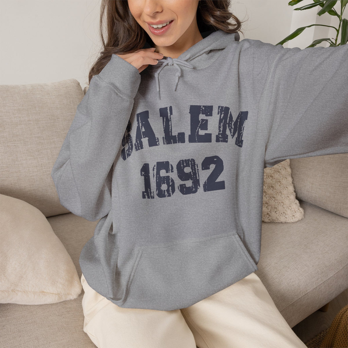 A woman wearing a heather grey pullover hoodie with 'SALEM 1692' printed in distressed navy blue letters across the chest, sitting on a beige couch.
