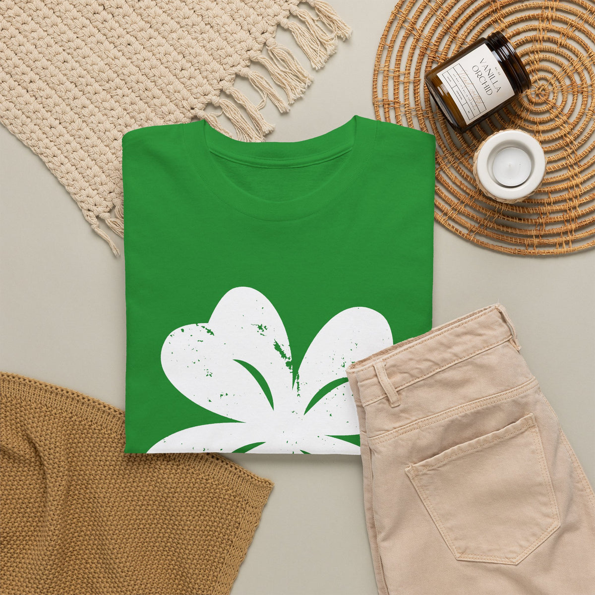 A green short-sleeve T-shirt with a large white four leaf clover graphic on the front, styled in a flat lay with beige pants, a woven rug, a candle jar, and a small white cup on a neutral background.