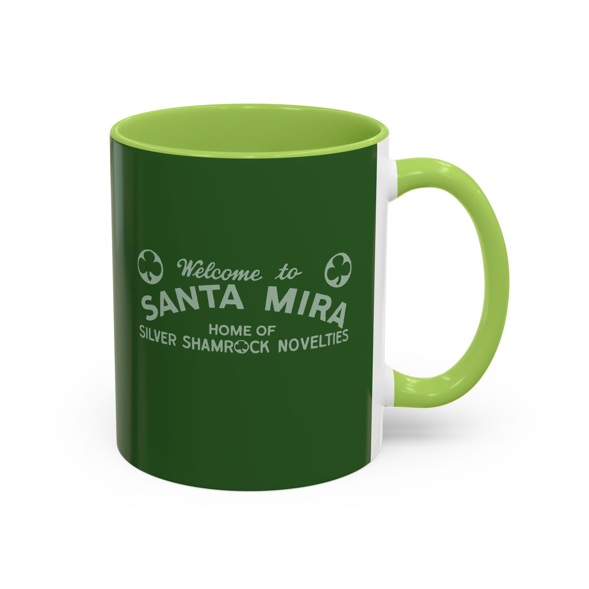 A ceramic mug with a dark green exterior and a light green handle and interior, featuring the white printed text 'Welcome to SANTA MIRA HOME OF SILVER SHAMROCK NOVELITIES' with shamrock icons.