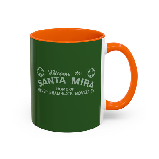A ceramic mug with a dark green exterior, bright orange handle and interior, and white printed text reading 'Welcome to SANTA MIRA HOME OF SILVER SHAMROCK NOVELITIES' on a white background.