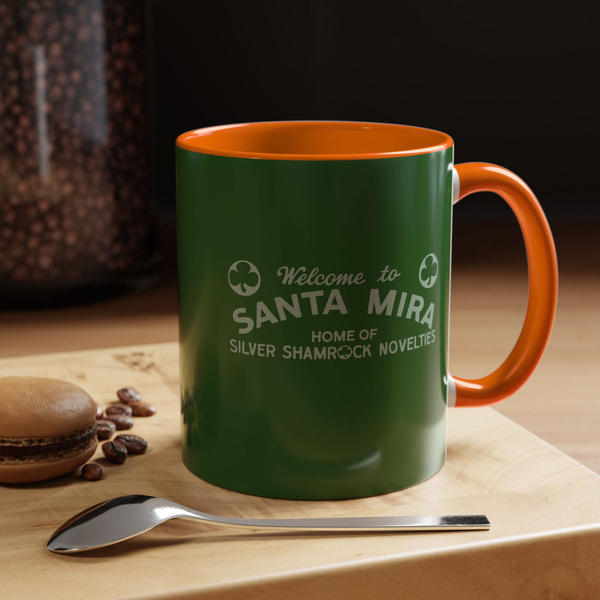 A green ceramic coffee mug with an orange handle and interior, printed with the text 'Welcome to SANTA MIRA HOME OF SILVER SHAMROCK NOVELTES' in white. The mug is shown on a wooden board next to a spoon and a macaron, with a dark background.