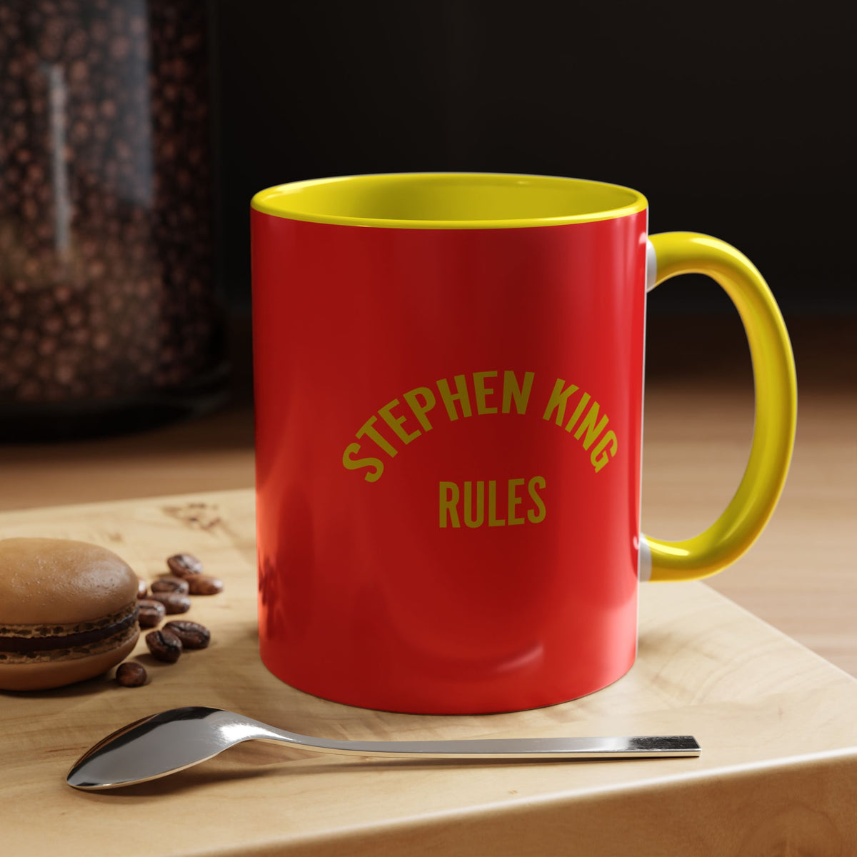 A red ceramic mug with a yellow handle and interior, printed with the text 'STEPHEN KING RULES' in yellow. The mug is placed on a wooden board next to a spoon and coffee beans, with a dark background.